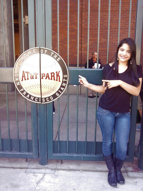 mulher do cabelo preto, pele branca, magra. Vestida de jeans, bola e camiseta preta apontando para uma placa do AT&T Park San Francisco